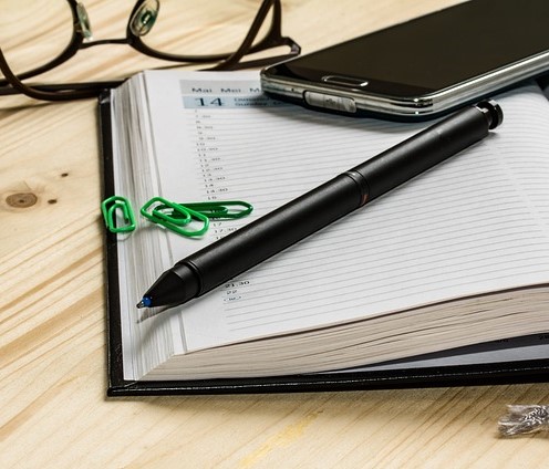 Kalenderbuch aufgeschlagen mit Kugelschreiber innenliegend, Handy und Büroklammern, Brille im Hintergrund