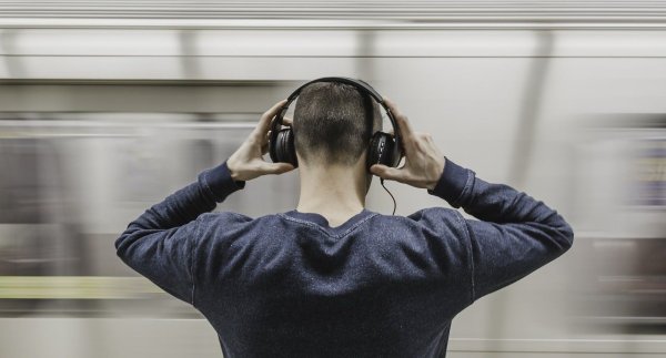Mann mit blauem Pullover, Rückenansicht, beide Hände am Kopf mit Kopfhörer, Zug fährt vorbei im Hintergrundd