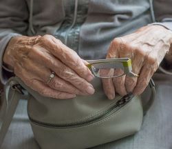 Hände einer Seniorin, die ihre Handtasche umfasst, gleichzeitig hält sie in der rechten Hand ihre Lesebrille